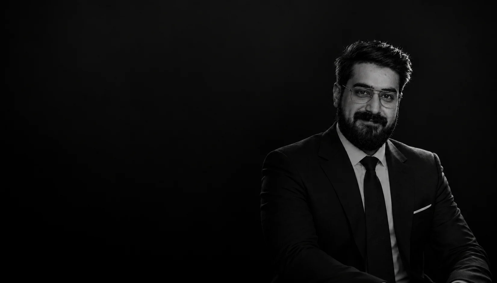 Ibraheem Hassan in a dark suit and tie, seated and lit from the right against a black background.